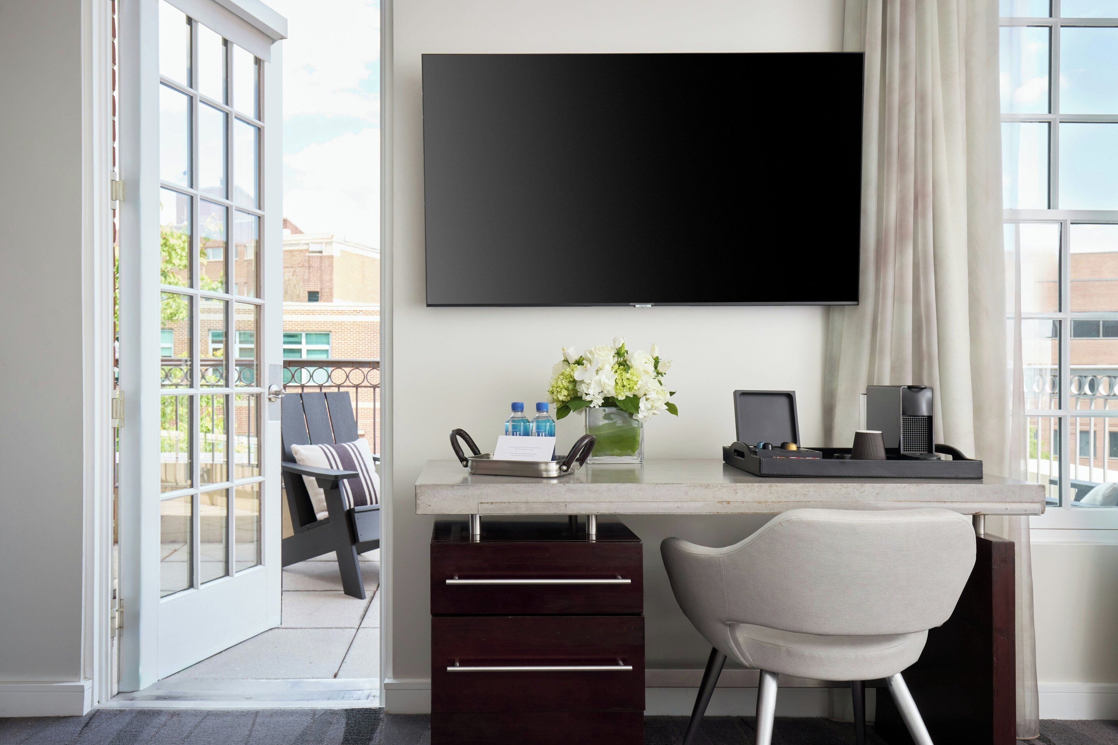 Ein modernes Hotelzimmer mit einem großen Flachbildfernseher an der Wand, einem Schreibtisch mit einem Laptop, einer Vase mit Blumen und einem Sessel.