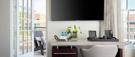 Ein modernes Hotelzimmer mit einem großen Flachbildfernseher an der Wand, einem Schreibtisch mit einem Laptop, einer Vase mit Blumen und einem Sessel.