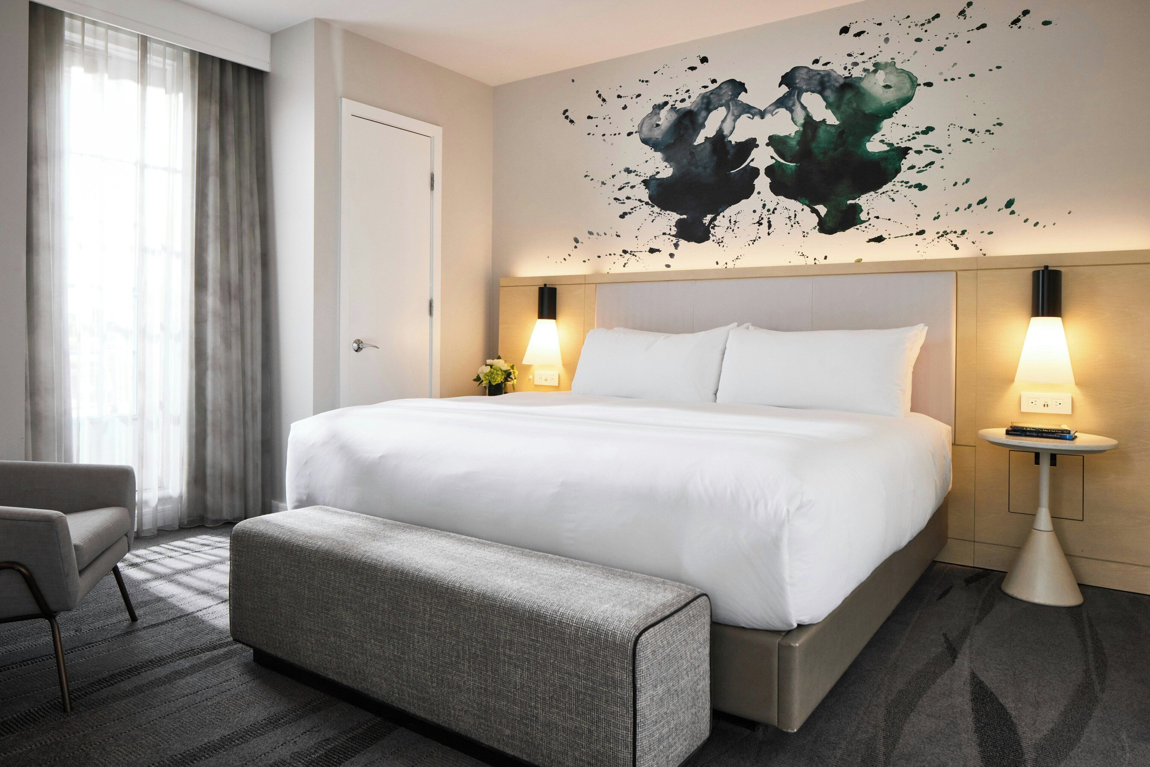 Una habitación de hotel con una cama grande, lámparas de noche, un banco, una obra de arte en la pared y una ventana con cortinas.