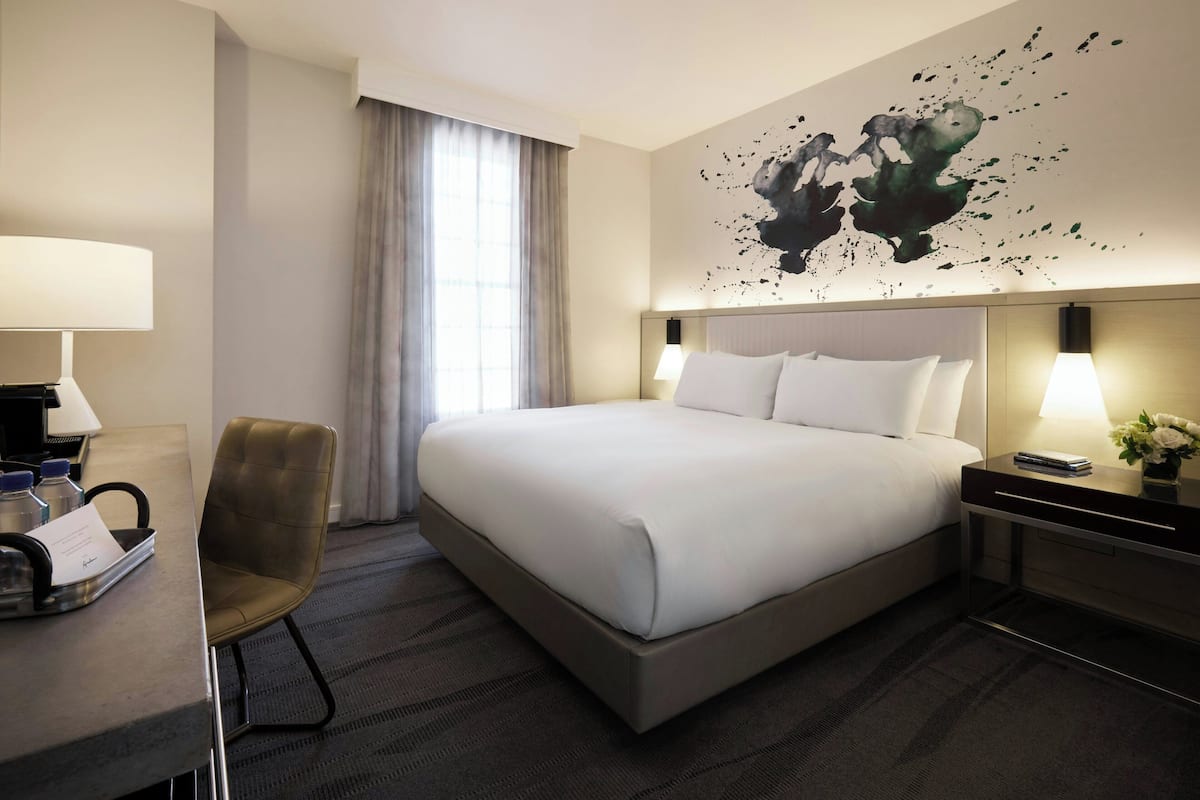 Quarto de hotel com uma cama grande, uma escrivaninha com cadeira, mesa de cabeceira com abajur e uma obra de arte na parede.