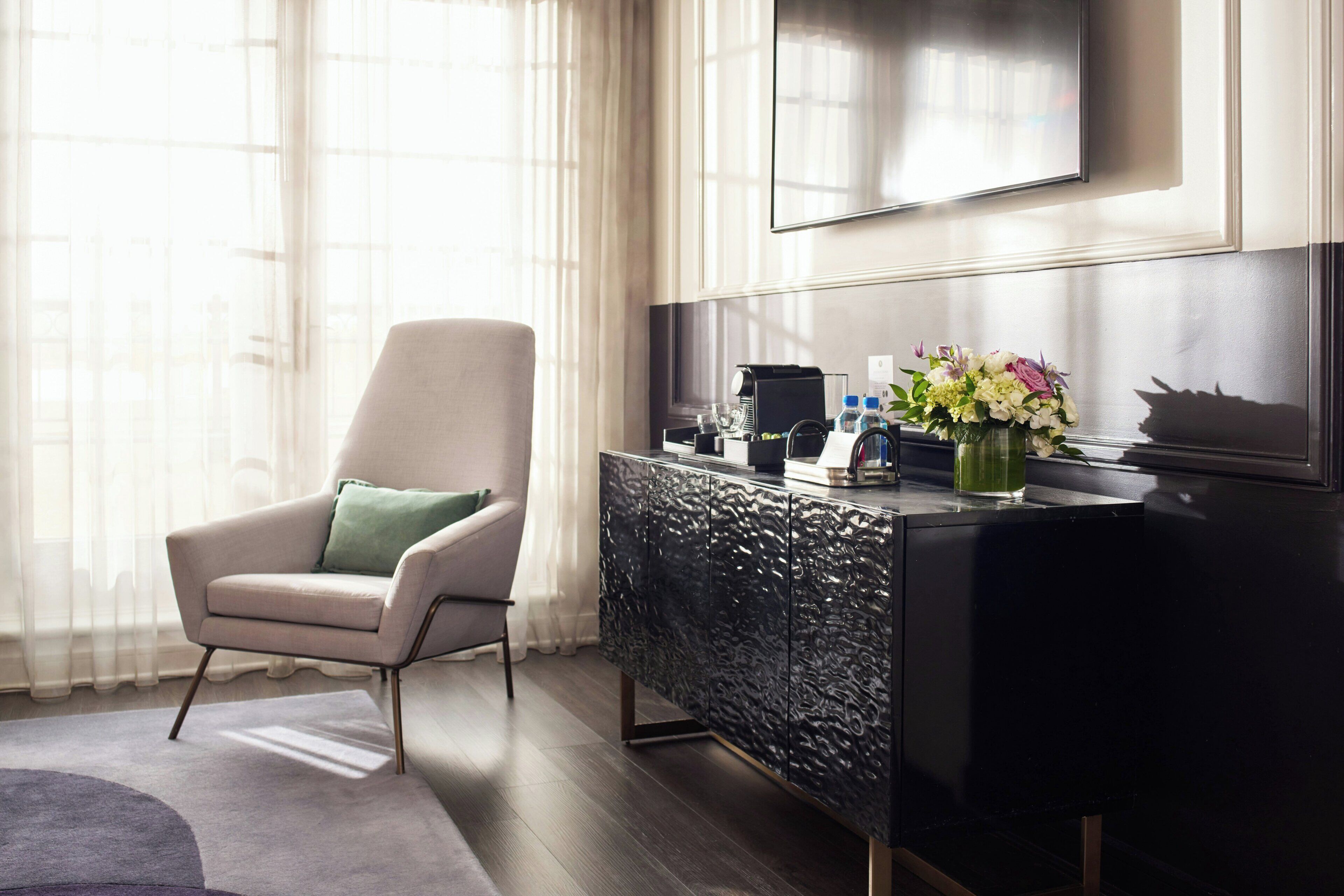 Une chambre d’hôtel moderne, dotée d’un grand téléviseur à écran plat fixé au mur, d’une console sombre au revêtement texturé, d’un fauteuil confortable agrémenté d’un coussin vert et d’un vase de fleurs posé sur la console.