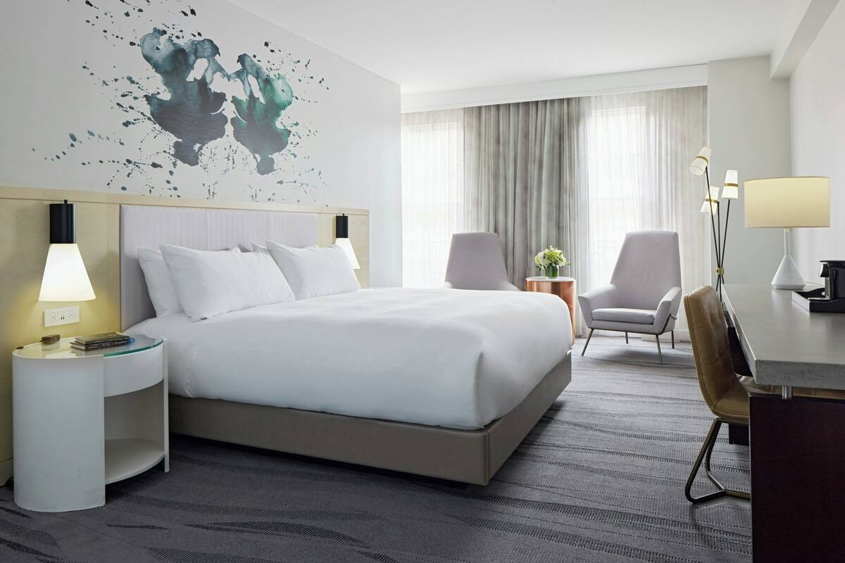 Quarto de hotel com uma cama grande, uma escrivaninha, duas cadeiras, um abajur e uma obra de arte na parede.