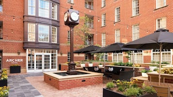 A courtyard with a clock, outdoor seating, and a brick building with a sign reading 'ARCHER HOTEL'.