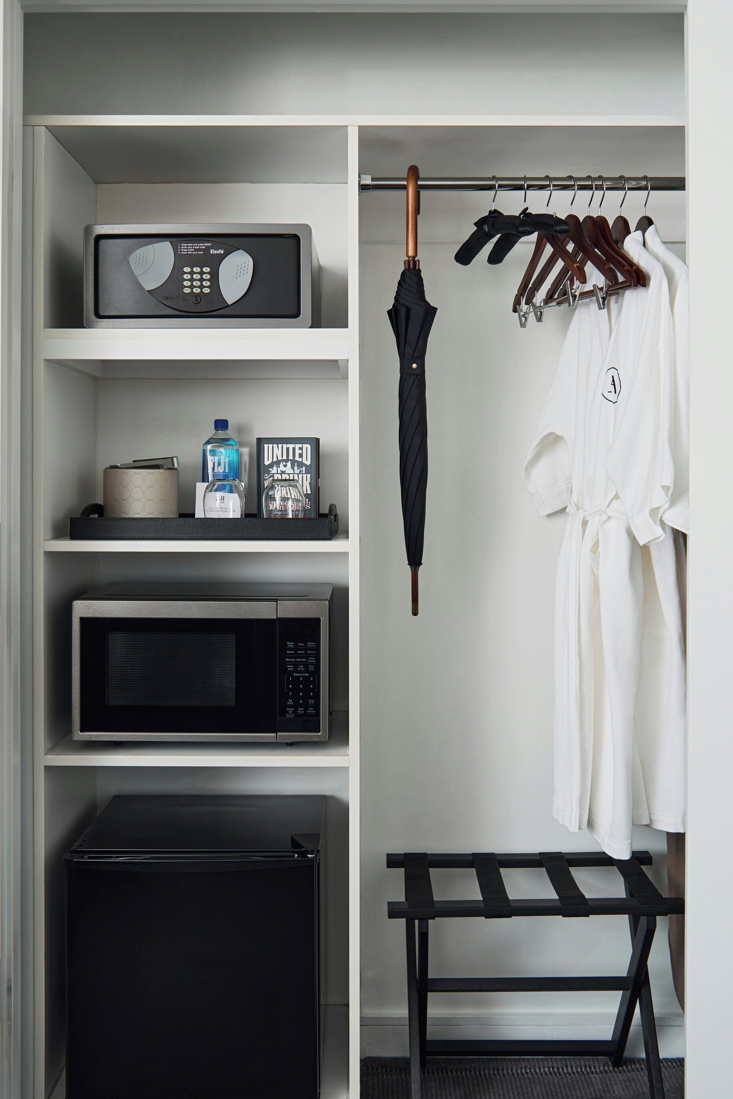 A closet with a microwave, a small refrigerator, a shelf with a water bottle, a book, and a black umbrella.