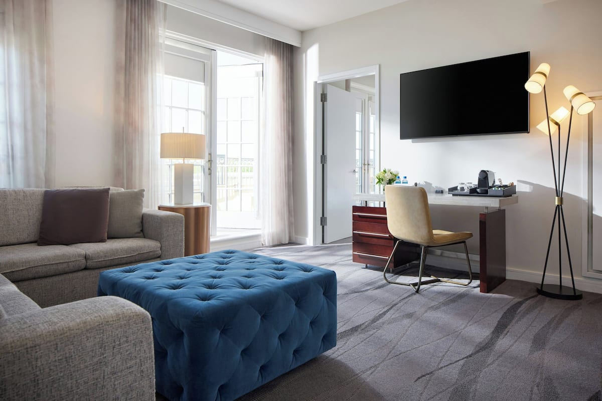 Quarto de hotel moderno com sofá, puf, escrivaninha, cadeira e televisão de tela plana.