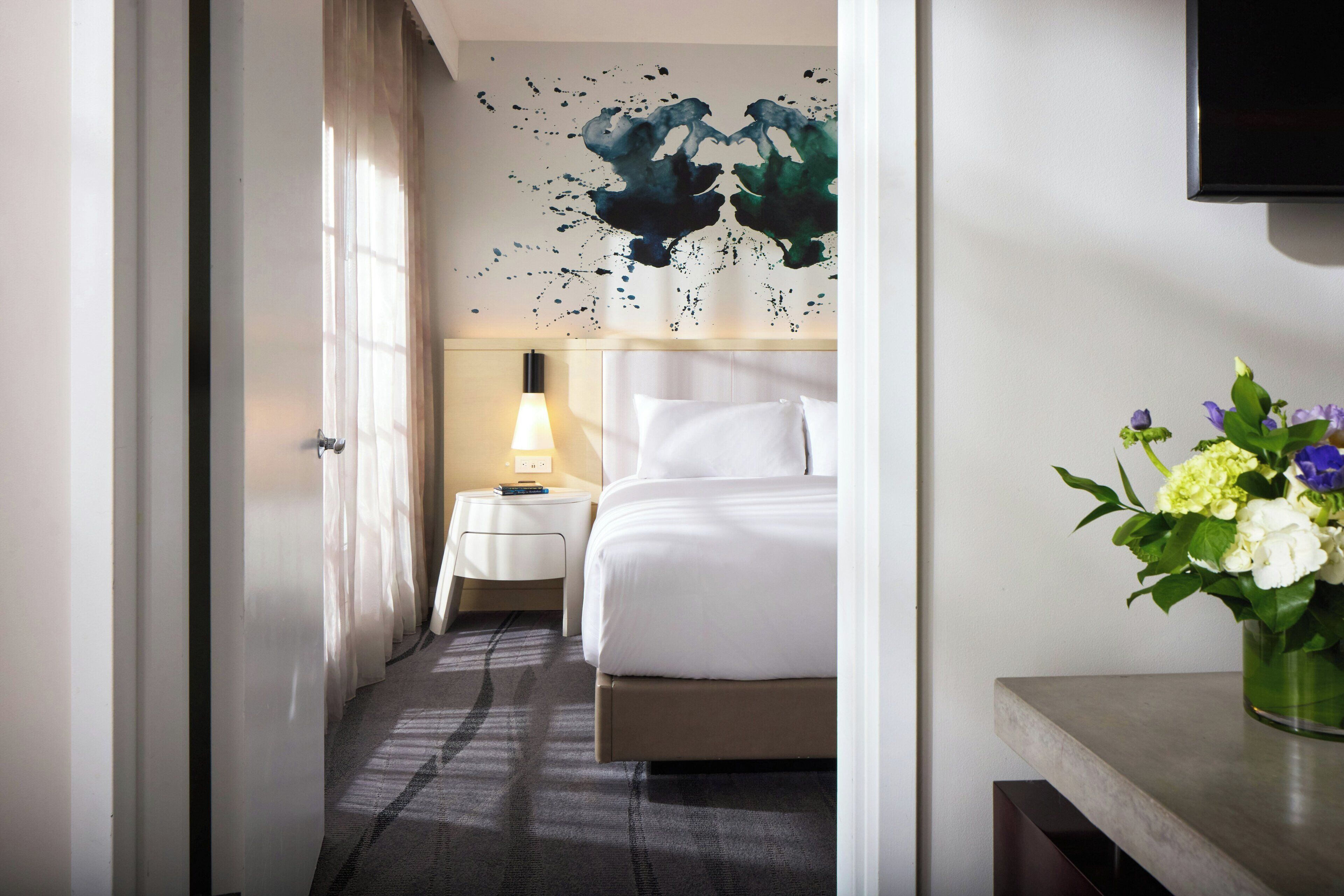 Ein Hotelzimmer mit Bett, Nachttisch mit Lampe, Wand-Fernseher und Blumen in einer Vase.