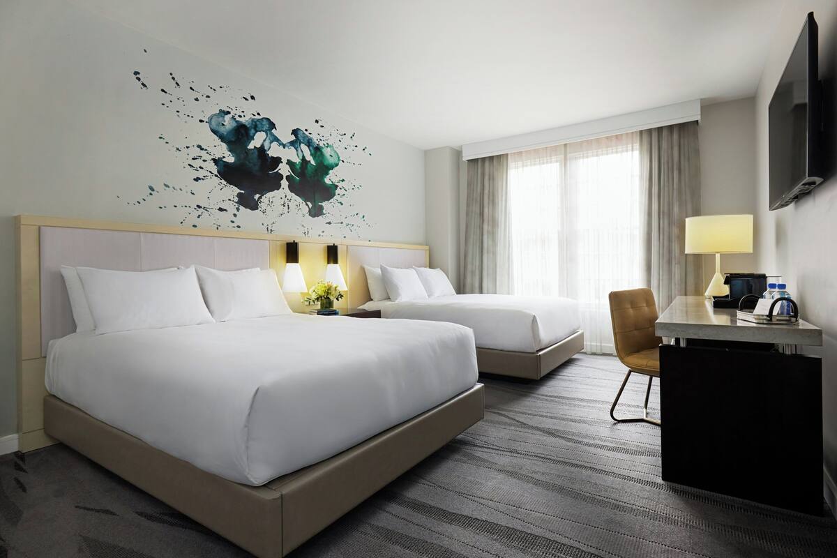 Quarto de hotel com duas camas, uma escrivaninha, uma cadeira, televisão e uma obra de arte na parede.