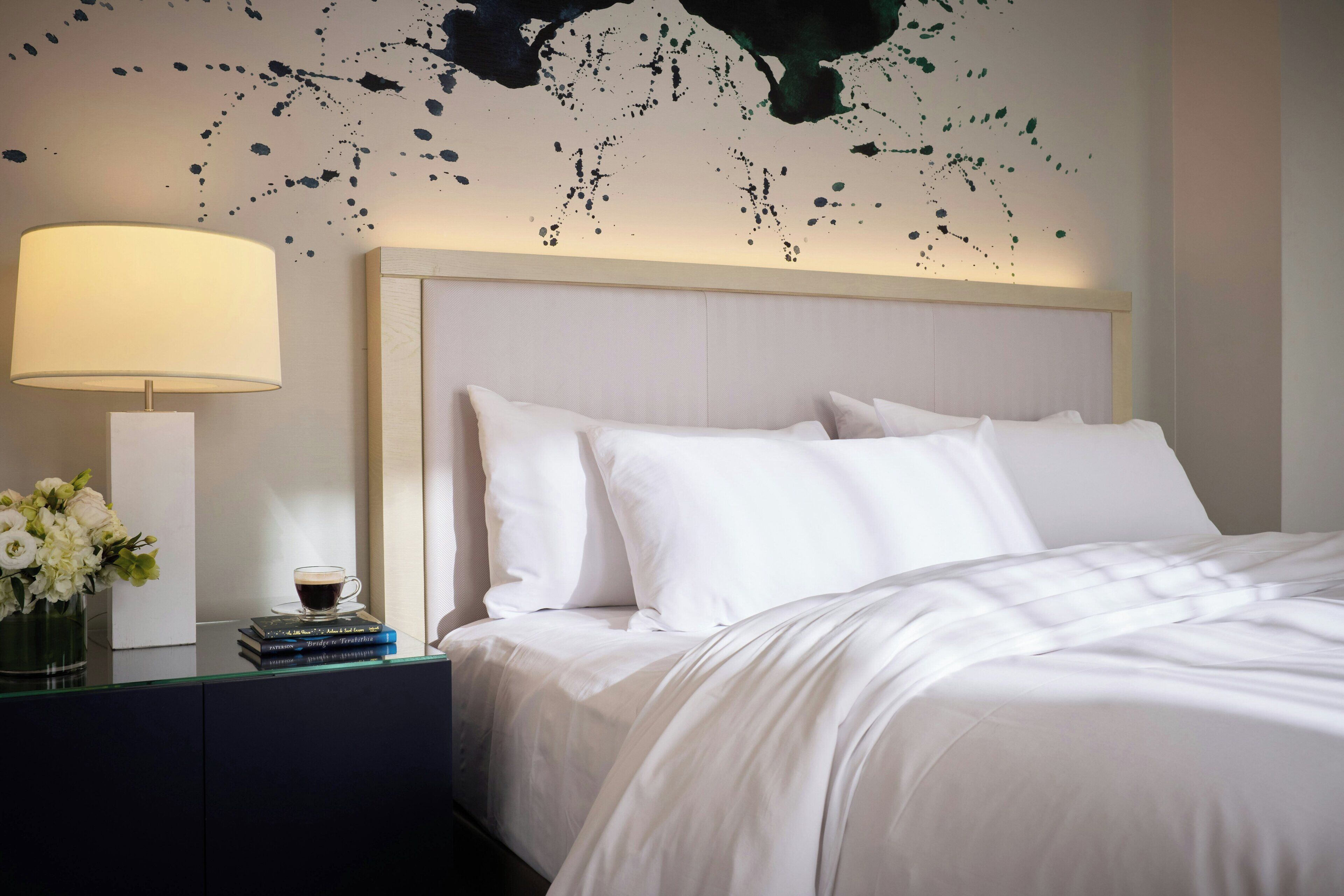 Ein ordentlich bezogenes Bett mit weißen Leinentüchern, ein Nachttisch mit Lampe und Büchern, und eine Wand mit einem einzigartigen, abstrakten Design.