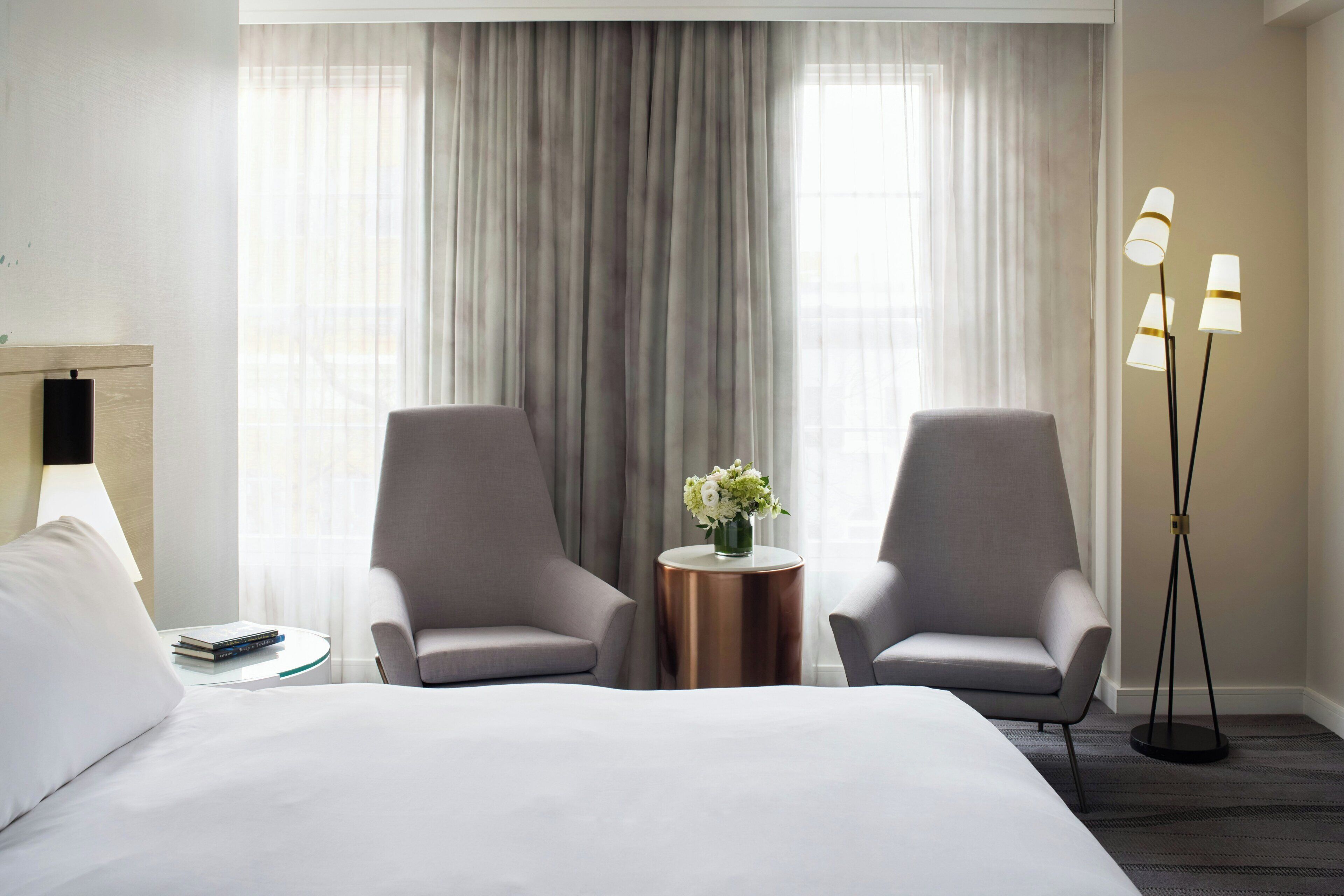 Quarto de hotel com uma cama, duas poltronas, uma mesa redonda com um vaso de flores e um abajur.