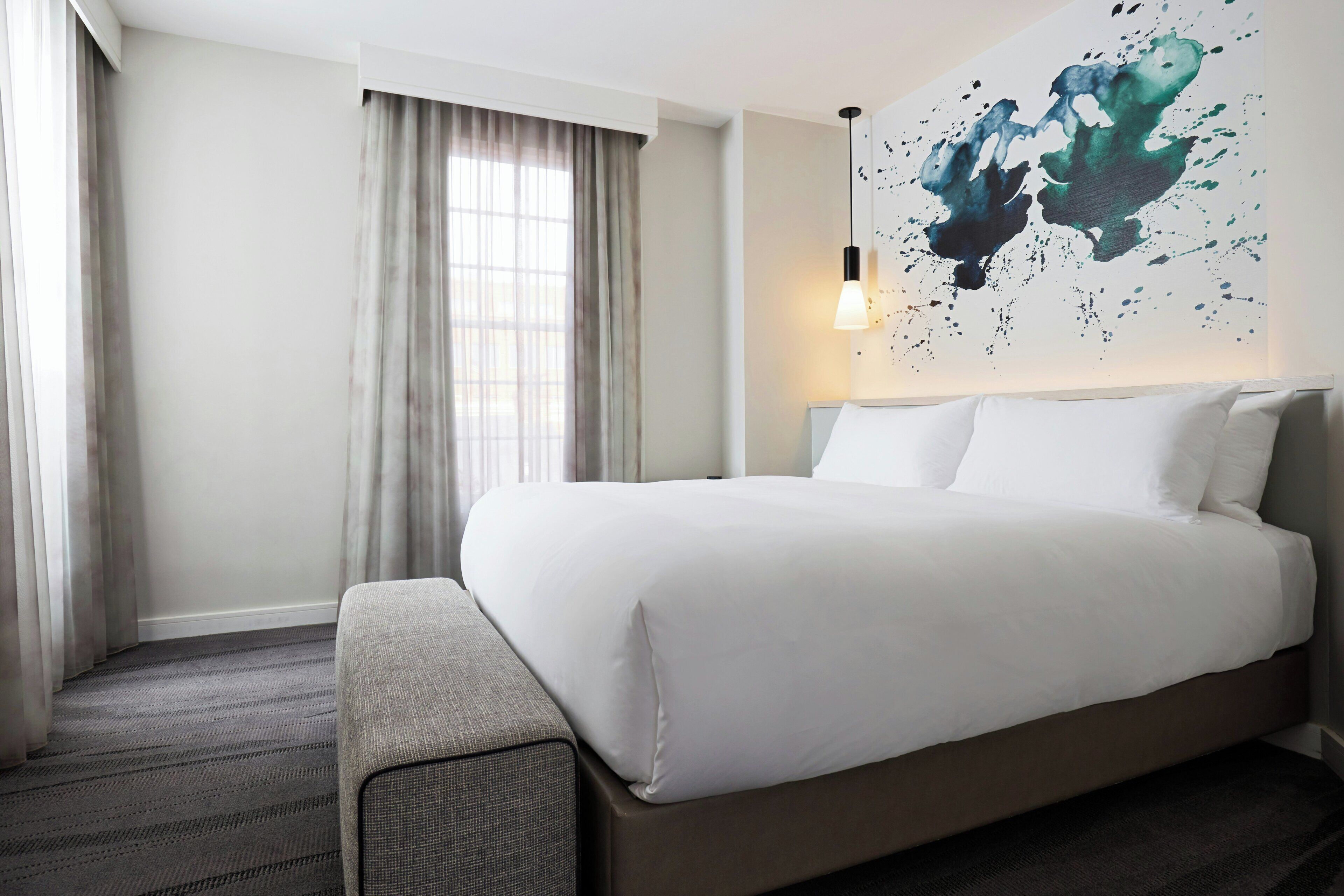 Una habitación de hotel con cama, una ventana con cortinas, una obra de arte en la pared y una lámpara colgante.