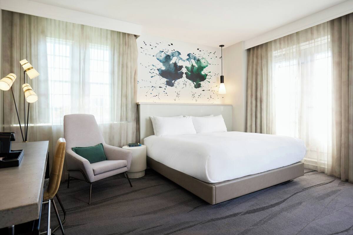 Quarto de hotel com uma cama grande, uma escrivaninha, uma cadeira e uma obra de arte na parede.