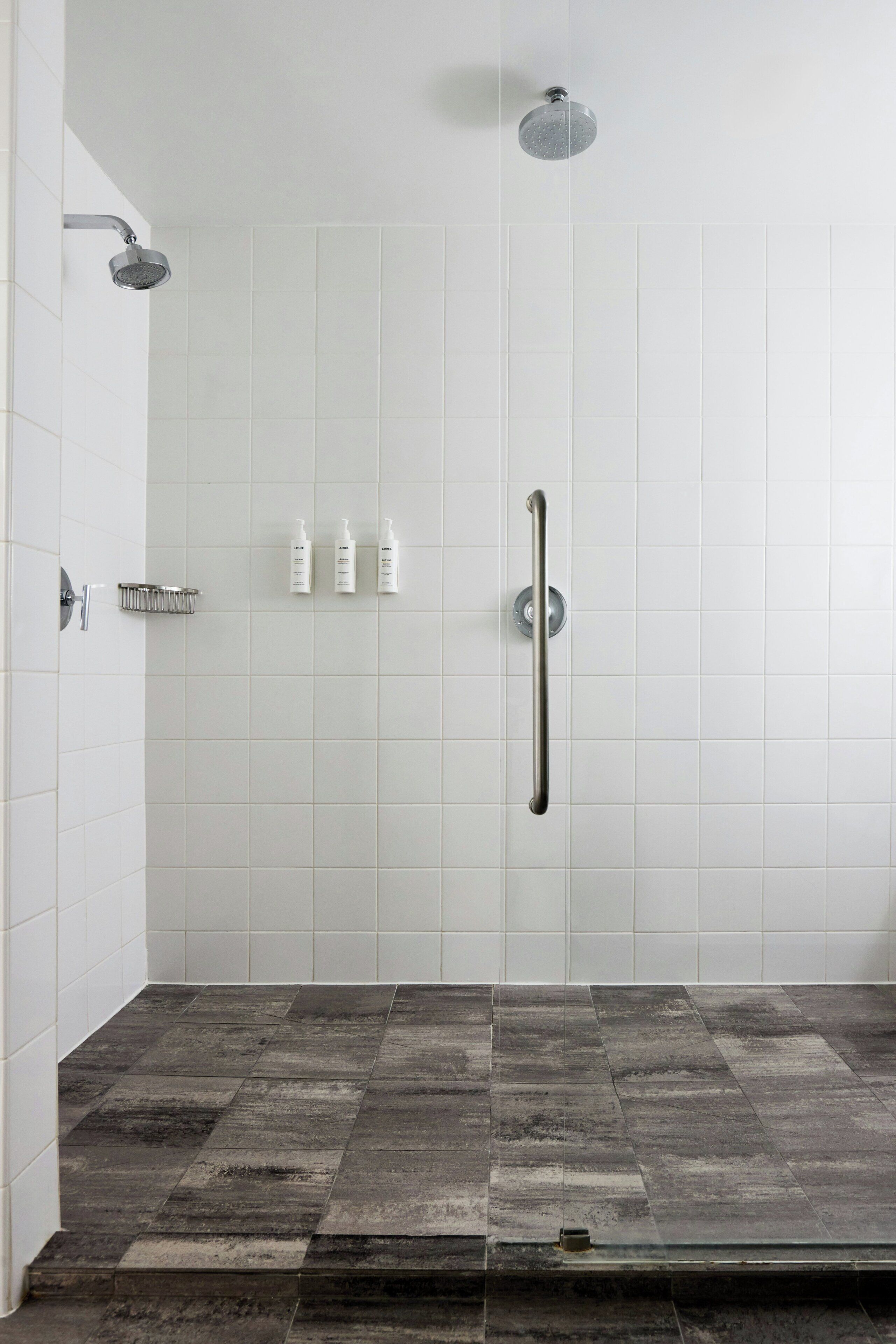 A shower with a handheld showerhead, a grab bar, and three bottles of shampoo on a wall-mounted shelf.