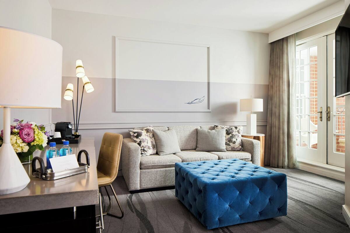Quarto de hotel com sofá, banquinho, escrivaninha, luminárias e uma janela grande com cortinas.