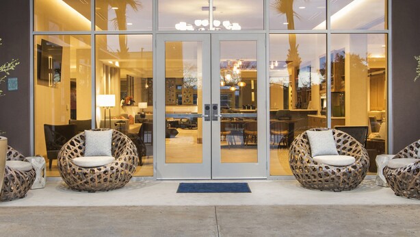 A hotel entrance with a glass door, two wicker chairs, and a reception area visible through the glass.