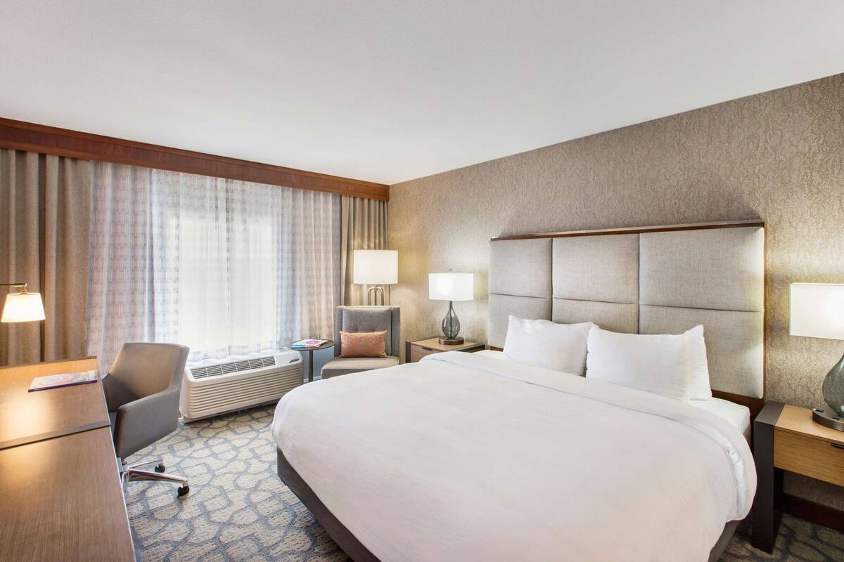 Ein Hotelzimmer mit einem großen Bett, einem Schreibtisch, einem Sessel, zwei Nachttischen mit Lampen, einer Klimaanlage und einem Fenster mit Vorhängen.
