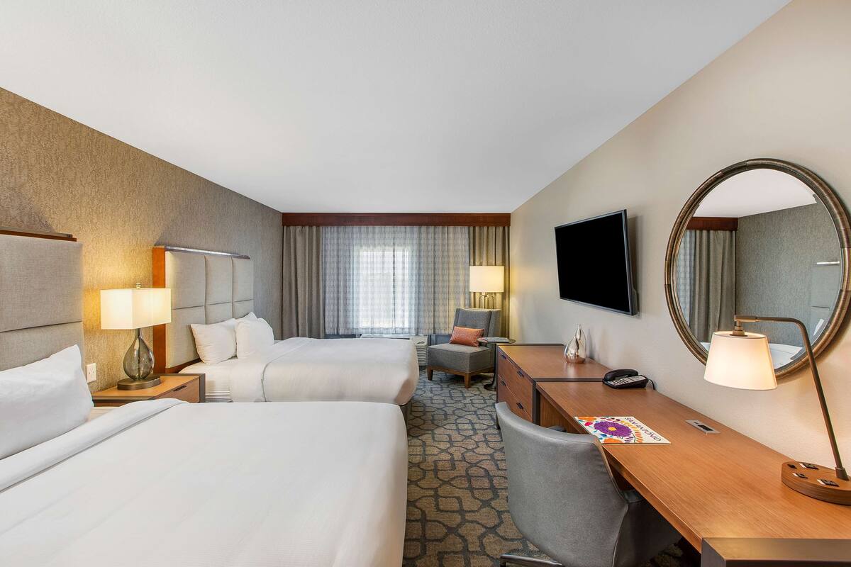 Ein Hotelzimmer mit zwei Betten, einem Schreibtisch, einem Stuhl, einem Fernseher, einem Spiegel und einem Fenster mit Vorhängen.