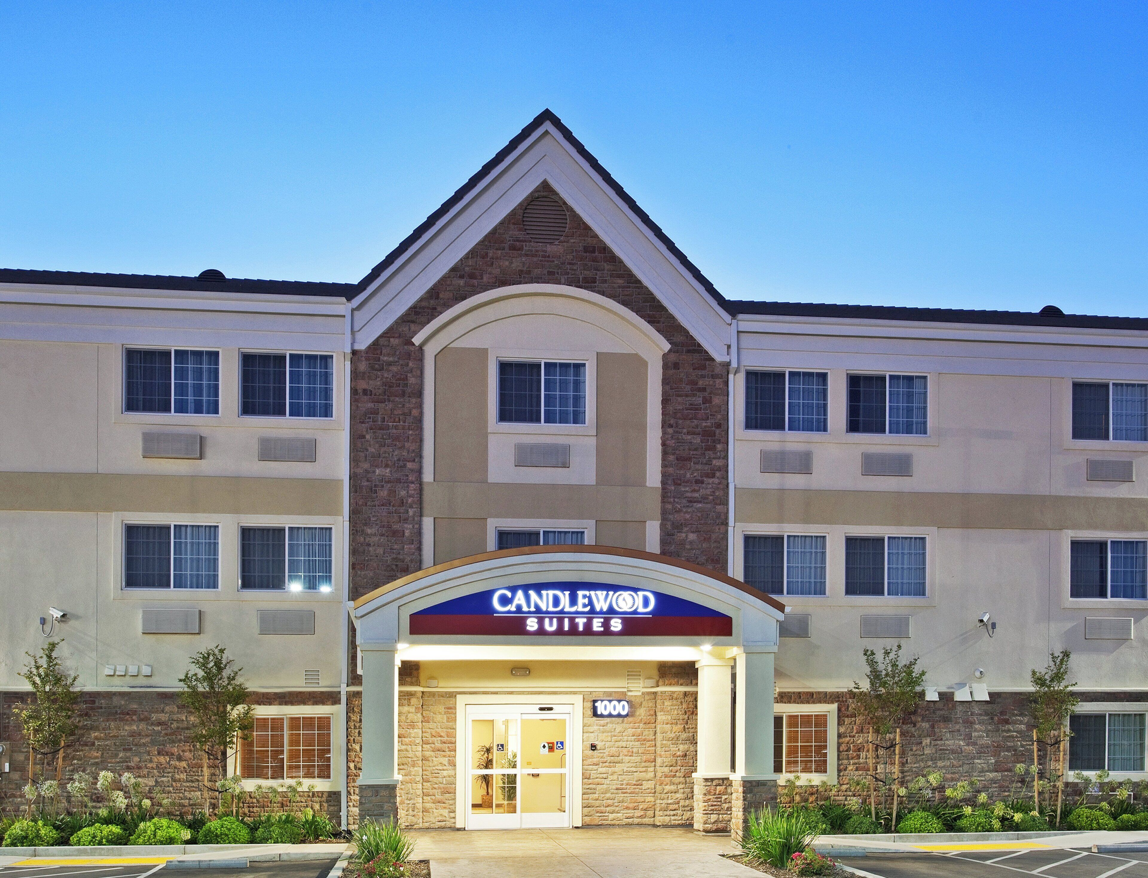 Een hotel met meerdere verdiepingen, een opvallende ingang, een bord met de tekst 'CANDLEWOOD SUITES' en het nummer '1000' als huisnummer.
