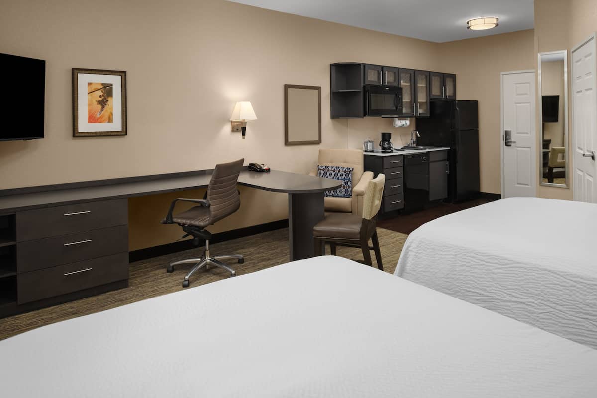 A hotel room with two beds, a desk with a chair, a small kitchenette, and a TV mounted on the wall.