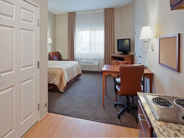 A hotel room with a bed, desk, chair, television, and a kitchen area with a stove.