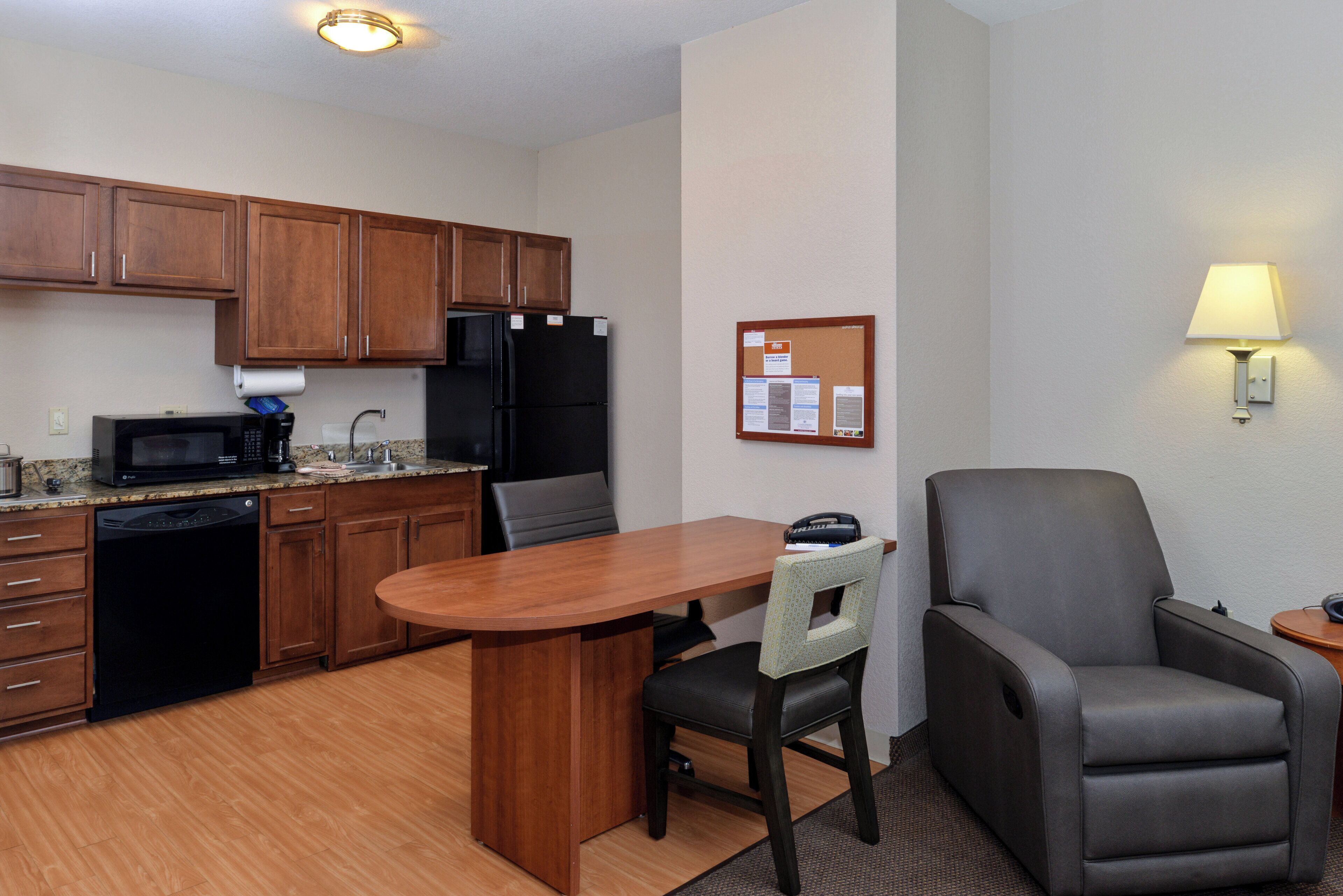 A compact kitchen with wooden cabinets, a black refrigerator, a microwave, and a coffee maker. There's a dining table with a chair, a bulletin board, and a lamp on the wall.