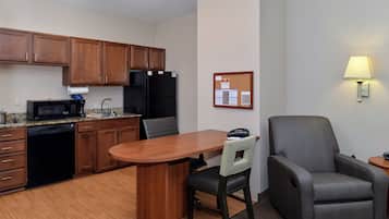 A compact kitchen with wooden cabinets, a black refrigerator, a microwave, and a coffee maker. There's a dining table with a chair, a bulletin board, and a lamp on the wall.