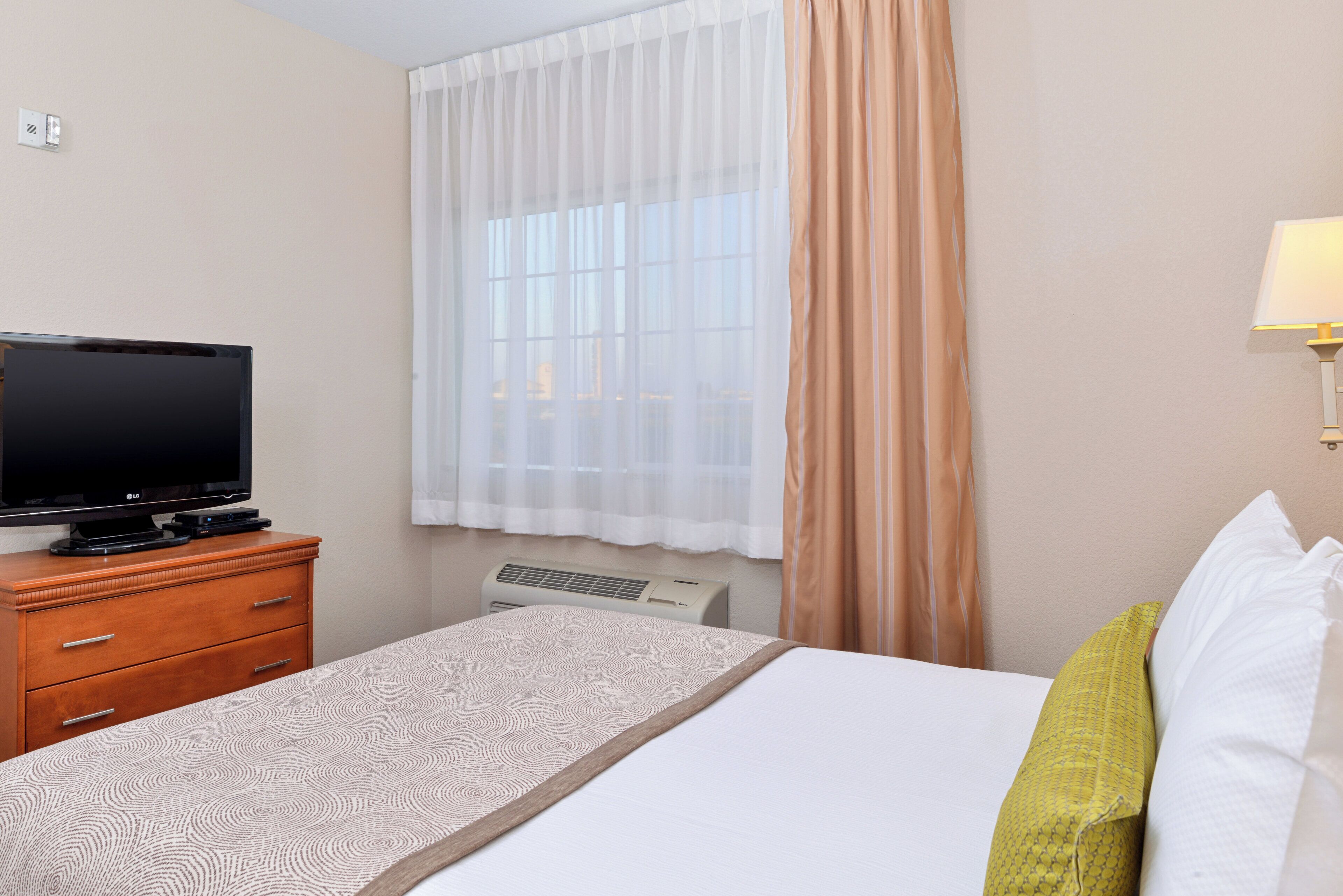 Habitación de hotel con cama, televisor sobre una cómoda de madera, ventana con cortinas ligeras y aire acondicionado de pared.