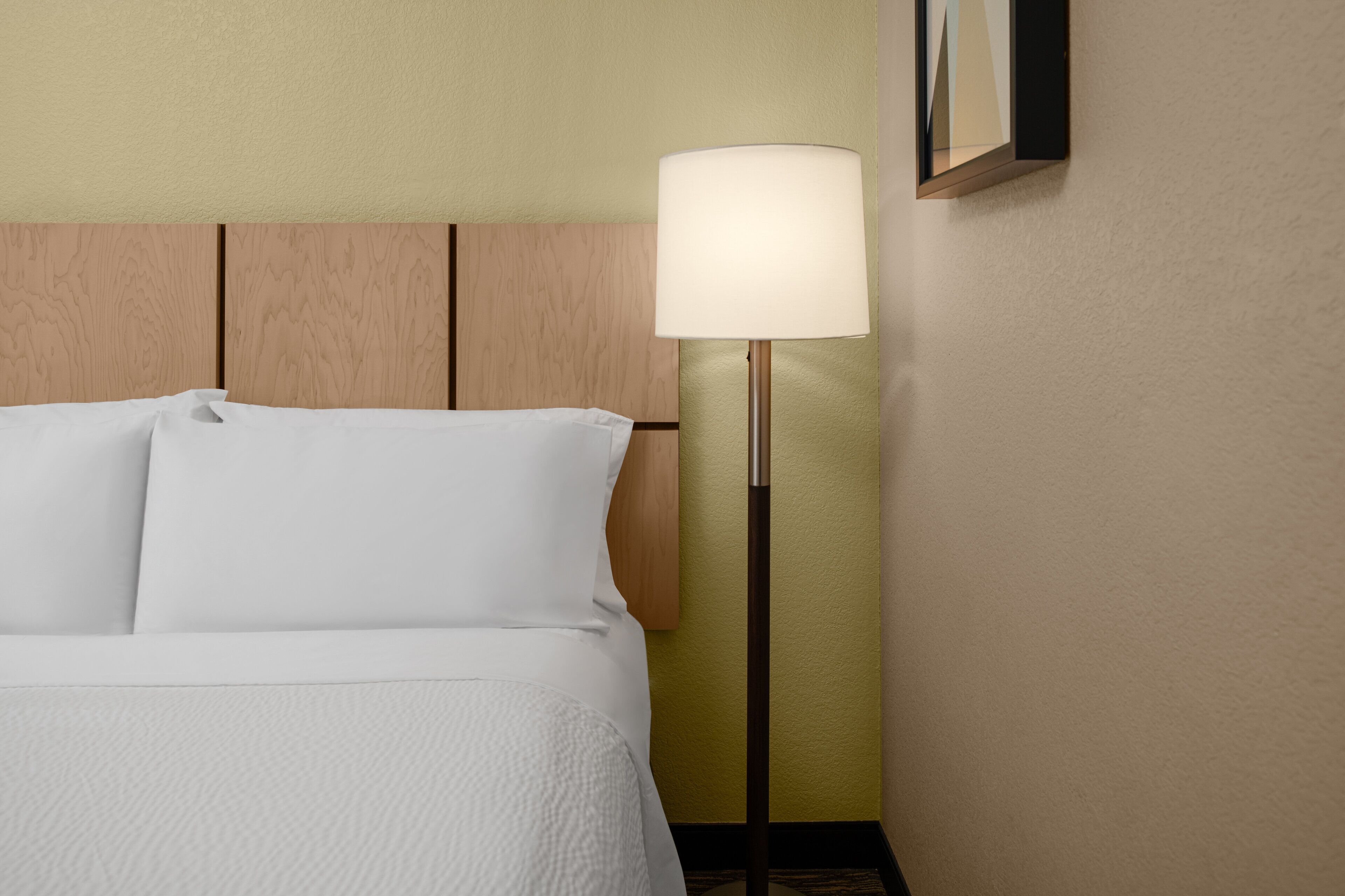 Quarto de hotel com cama, luminária e porta-retrato fixado na parede.