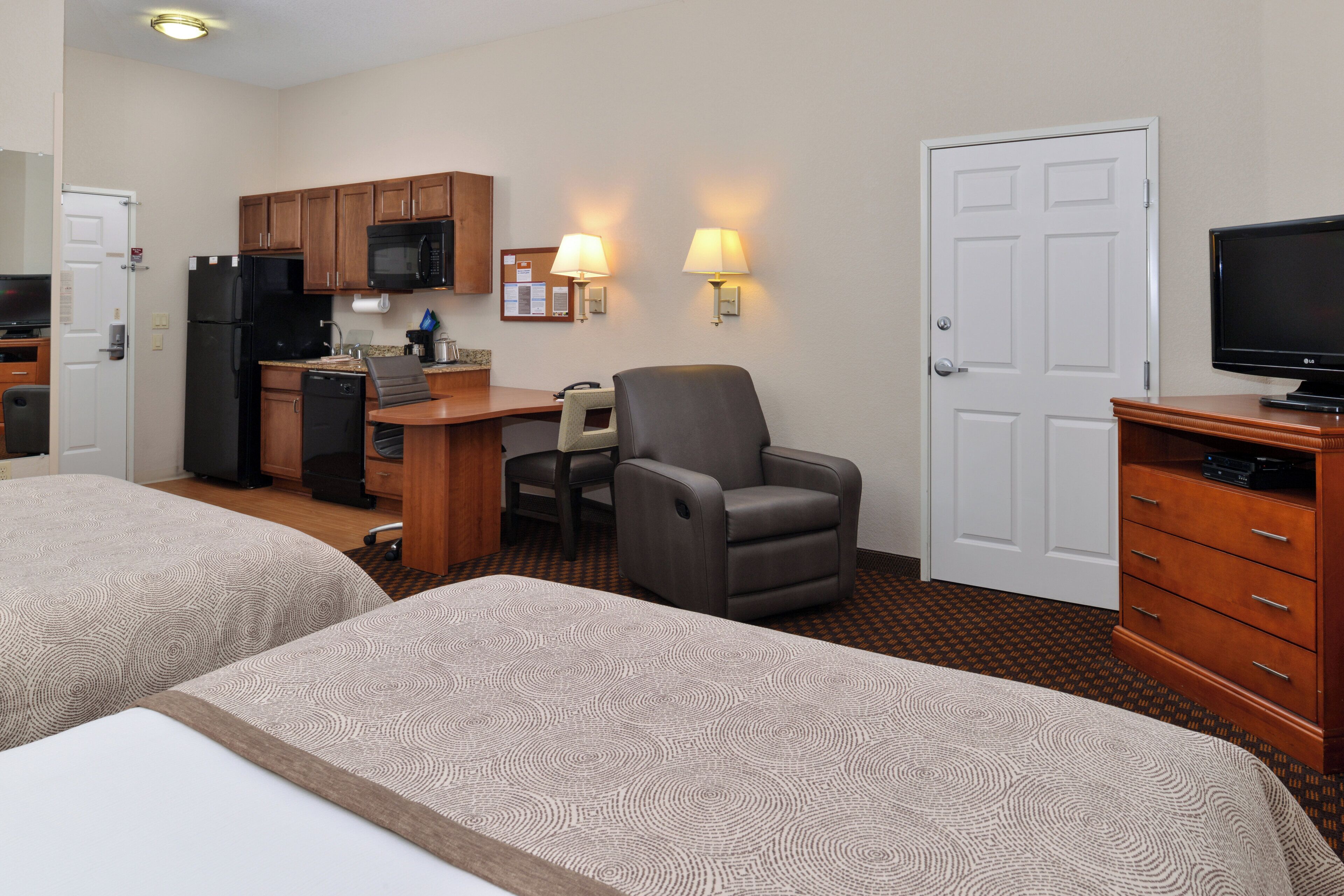 A hotel room with two beds, a kitchenette, a desk with a chair, a television, and a door.