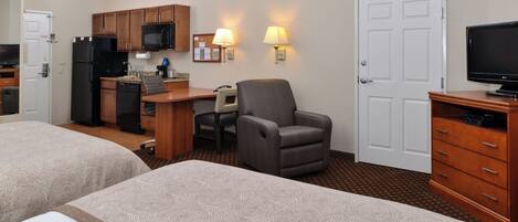 A hotel room with two beds, a kitchenette, a desk with a chair, a television, and a door.