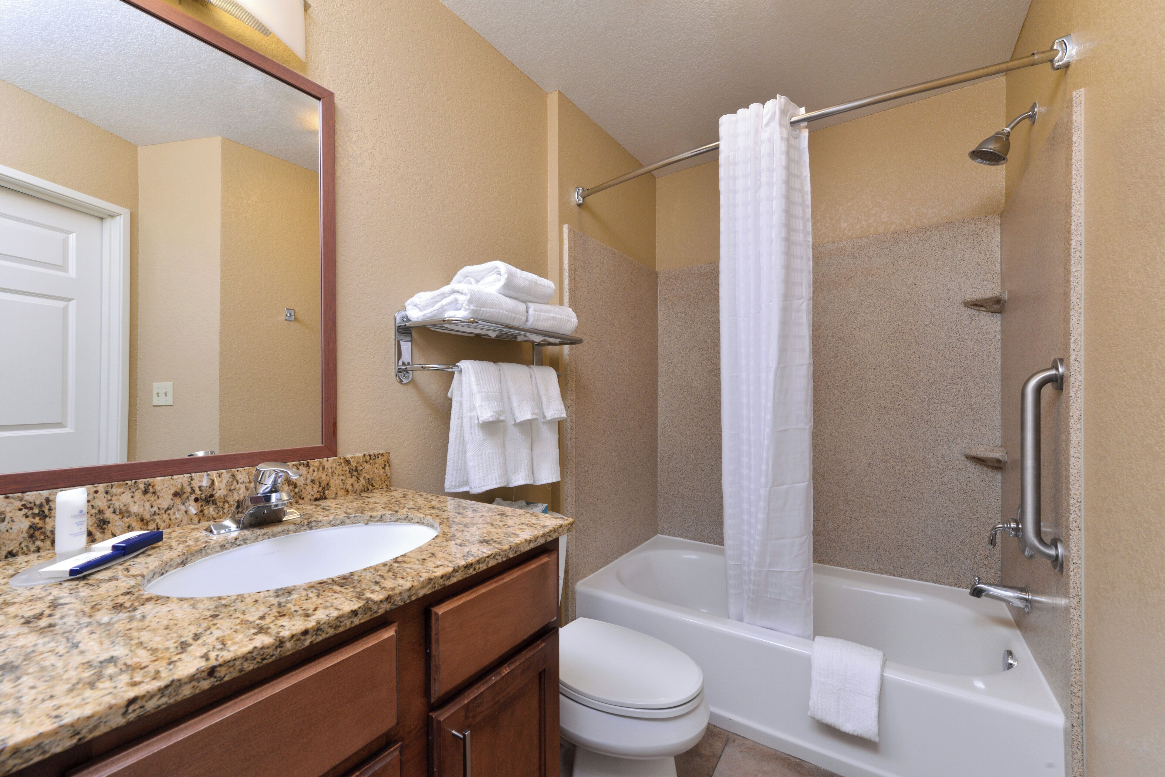 A bathroom with a granite countertop sink, a white toilet, a bathtub with a shower curtain, and a towel rack with folded towels.