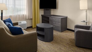 A hotel room with a sofa, a round table, a TV on a stand, a lamp, and a window with curtains.
