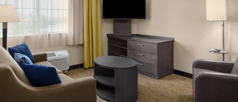 A hotel room with a sofa, a round table, a TV on a stand, a lamp, and a window with curtains.