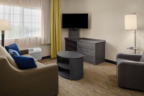 Une chambre d’hôtel avec un canapé, une table ronde, un téléviseur posé sur un meuble, une lampe et une fenêtre avec des rideaux.