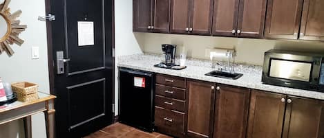 A compact kitchen with dark wooden cabinets, a granite countertop, a black refrigerator, a microwave, and a coffee maker.