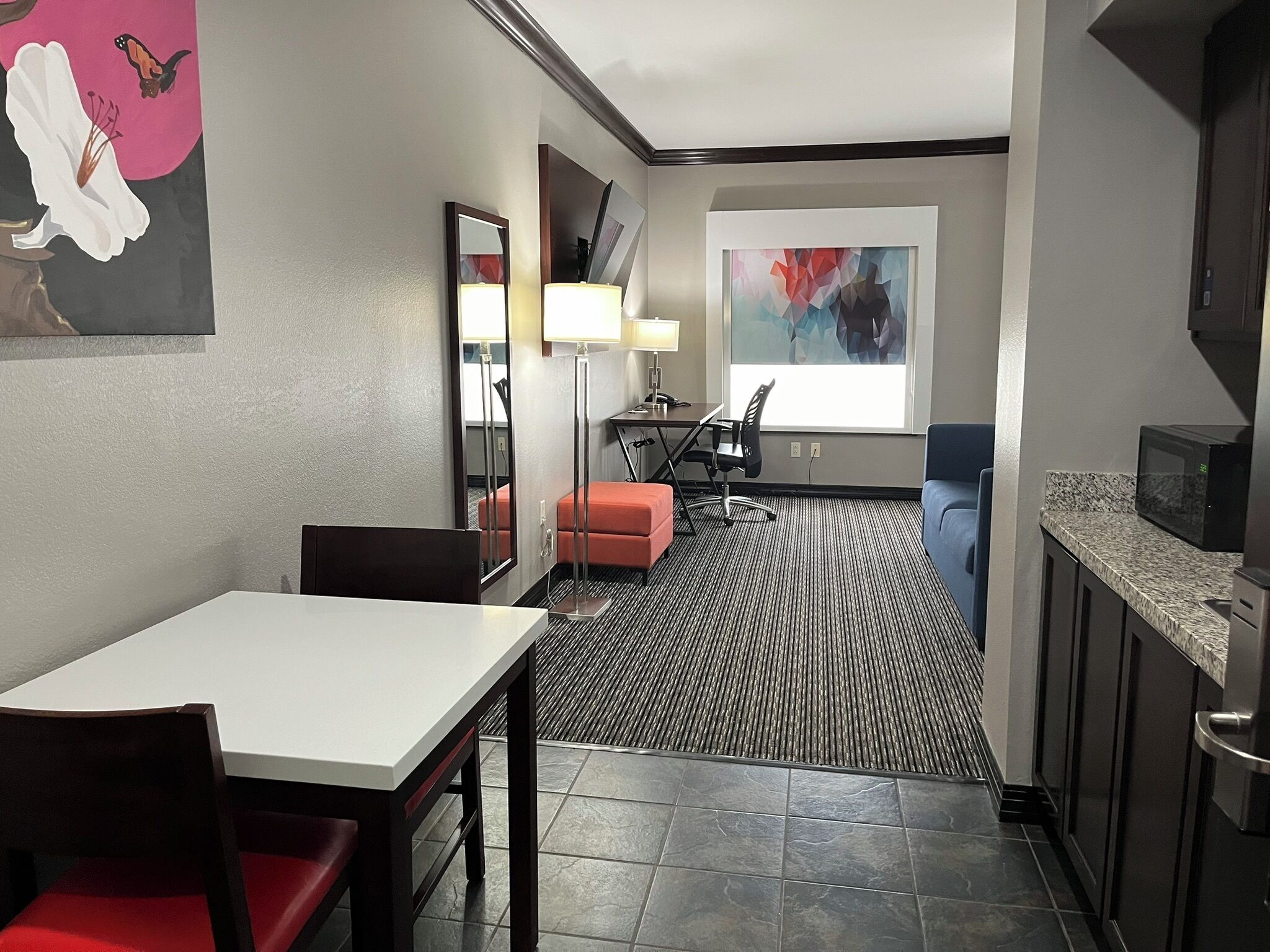 A hotel room with a dining table, chairs, a sofa, a small kitchen area with a microwave, and a large window with artwork.