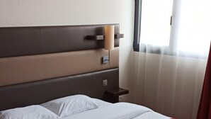 A neatly made bed with white linens and a red blanket, a bedside table with a lamp, and a window with curtains. - Résidence Hôtelière des Ondes (Saint-Lys)