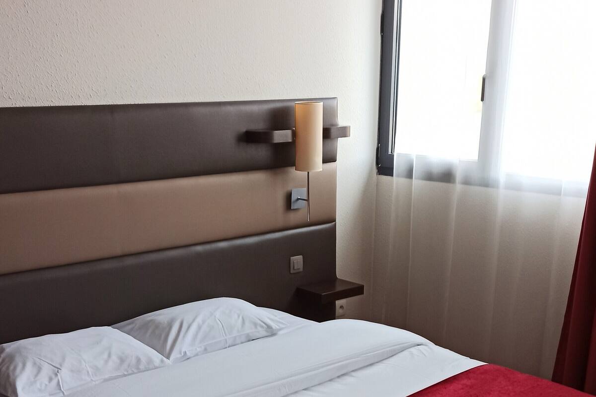 Een net opgemaakt bed met witte lakens en een rode deken, een nachtkastje met een lamp, en een raam met gordijnen.