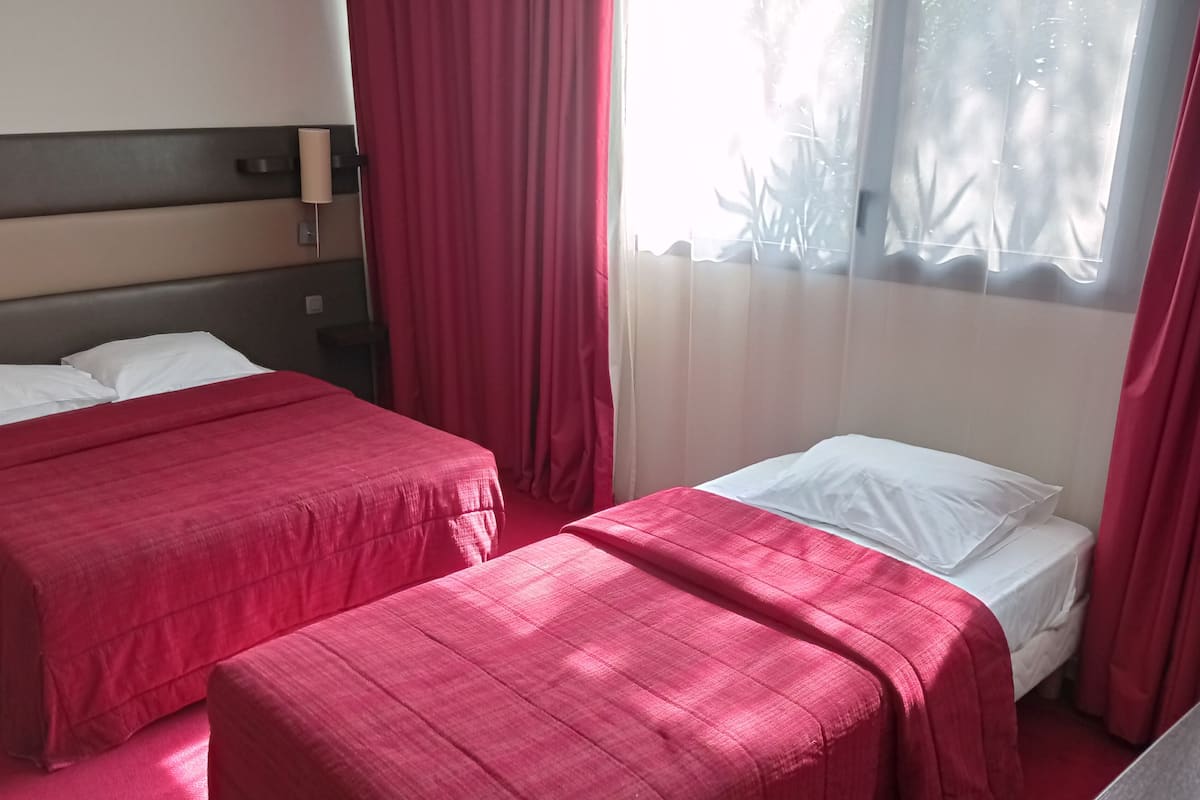 Een hotelkamer met twee bedden, elk met een rode sprei en witte kussens. Er is een nachtkastje met een lamp, en de kamer heeft een raam met rode gordijnen.