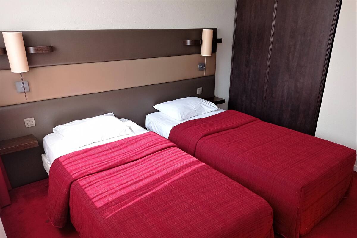 Een hotelkamer met twee bedden, elk met rode beddengoed en witte kussens. Er staan wandlampen op het hoofdeinde en een houten nachtkastje naast de bedden.
