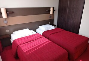 Une chambre d’hôtel avec deux lits, chacun doté de draps rouges et d’oreillers blancs. Des lampes murales sont fixées au-dessus de la tête de lit, et une table de chevet en bois se trouve à côté de chaque lit.