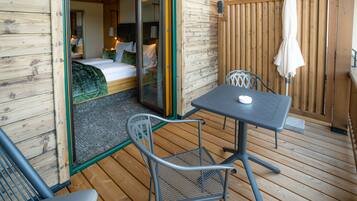 Eine Holzterrasse mit Tisch und Stühlen, eine Schiebeglastür, die zu einem Schlafzimmer mit Bett und Nachttisch führt.