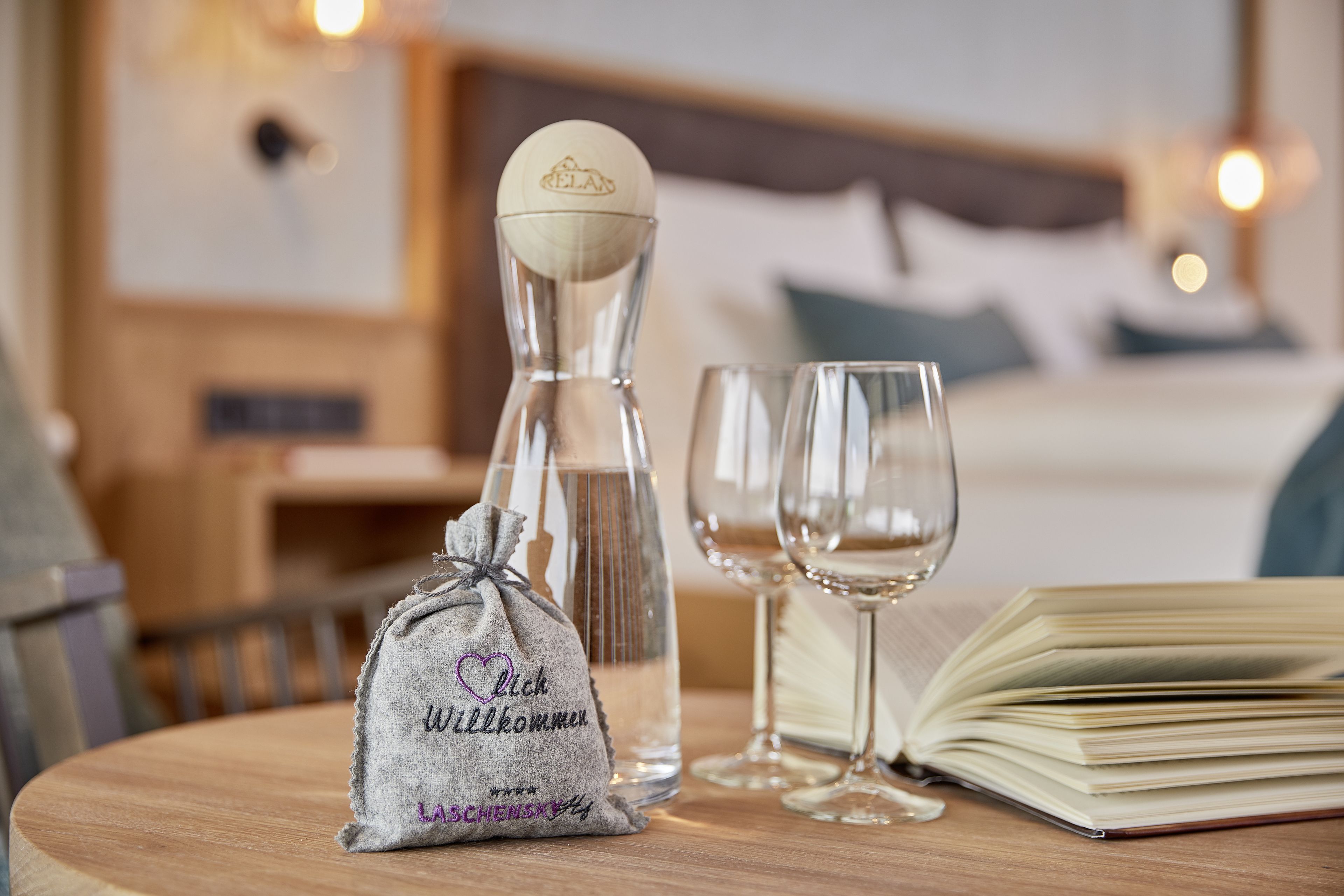 A small gift bag with a heart and text, a bottle, two wine glasses, and an open book on a wooden table in a hotel room.
