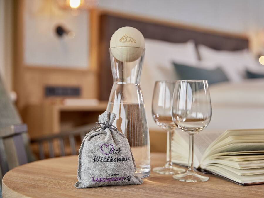 A small gift bag with a heart and text, a bottle, two wine glasses, and an open book on a wooden table in a hotel room.