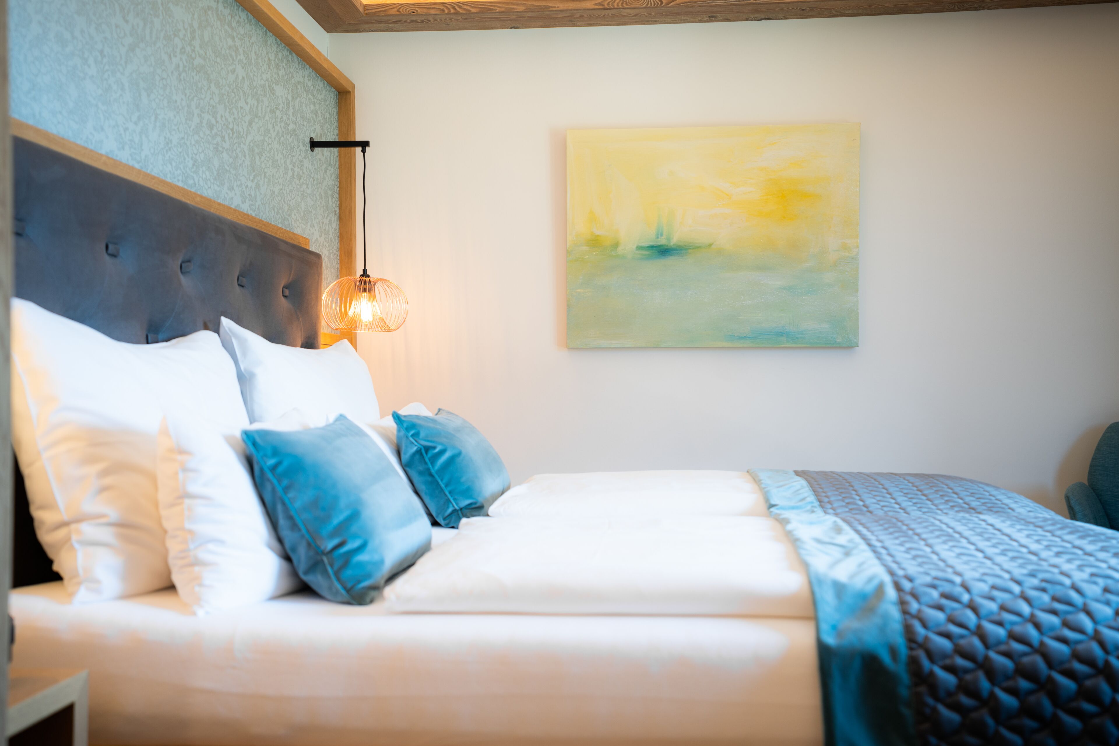 A neatly made bed with white and blue bedding, a wooden headboard, and a wall-mounted abstract painting.