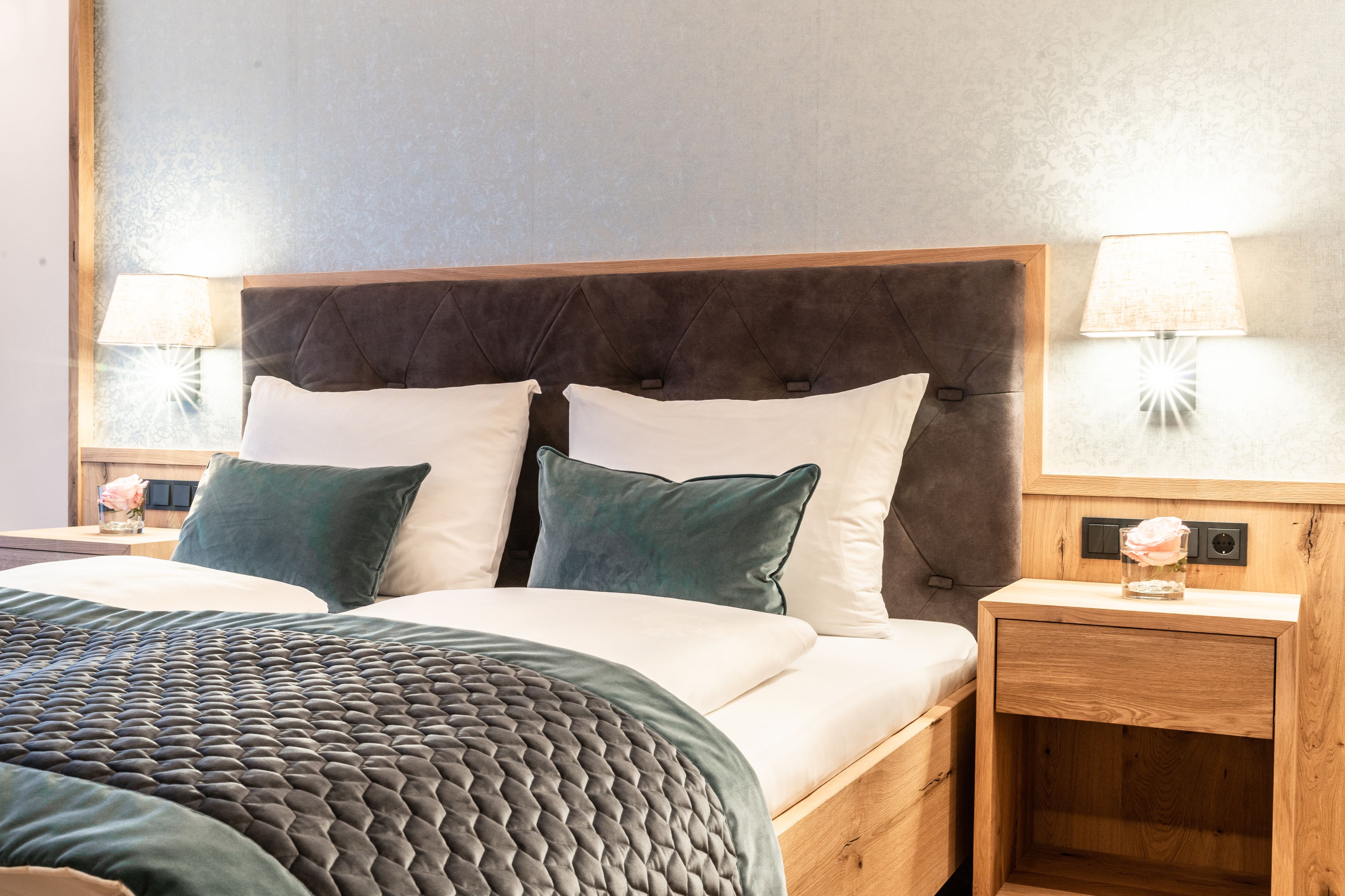 Una cama bien hecha con un cabecero oscuro, dos lámparas de noche y una mesita de madera con un florero.