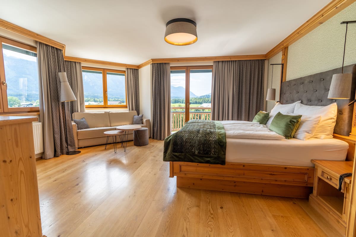 Un dormitorio amplio con una cama grande, un sofá, una mesita y vistas a montañas desde las ventanas.