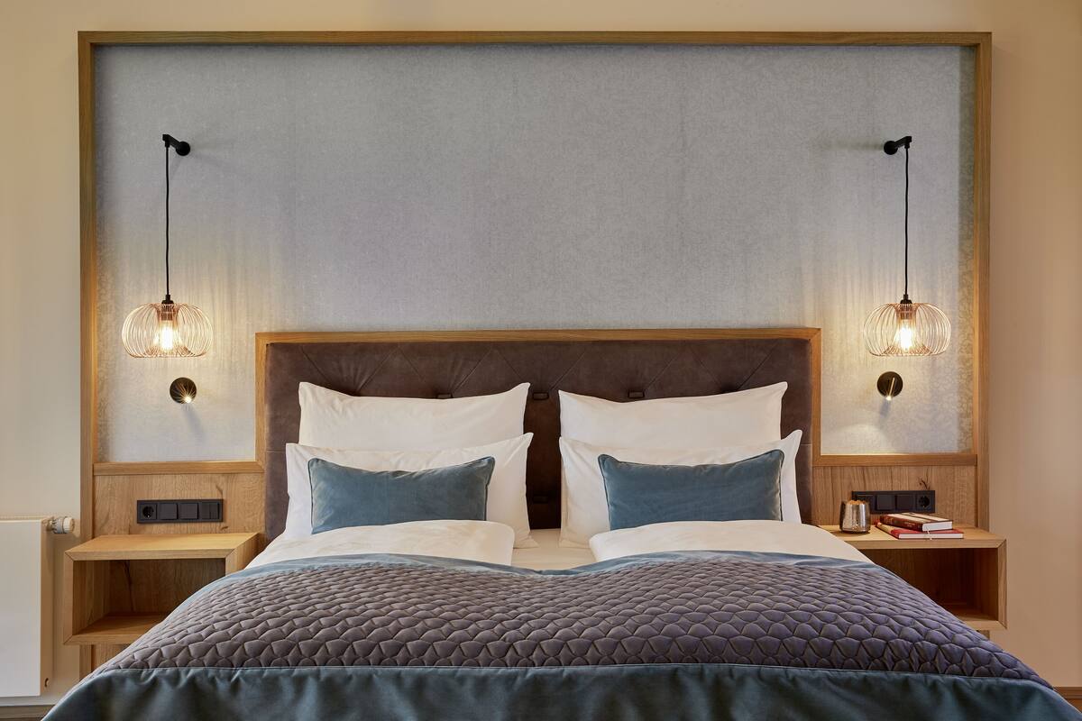 Una cama bien tendida con un cabecero acolchado, dos mesitas de noche con lámparas, y una lámpara de pared.