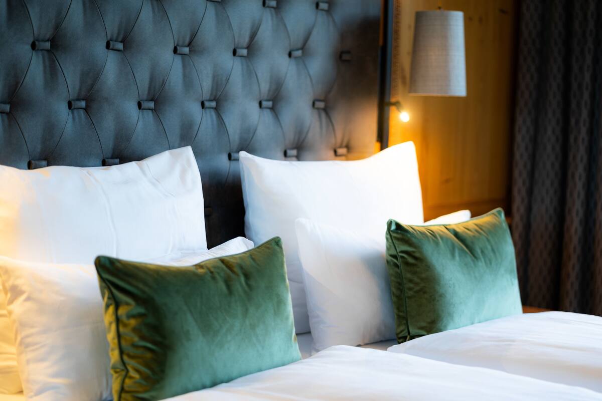 Una cama con ropa de cama blanca, almohadas verdes y un cabecero capitonado, con una lámpara en la pared.