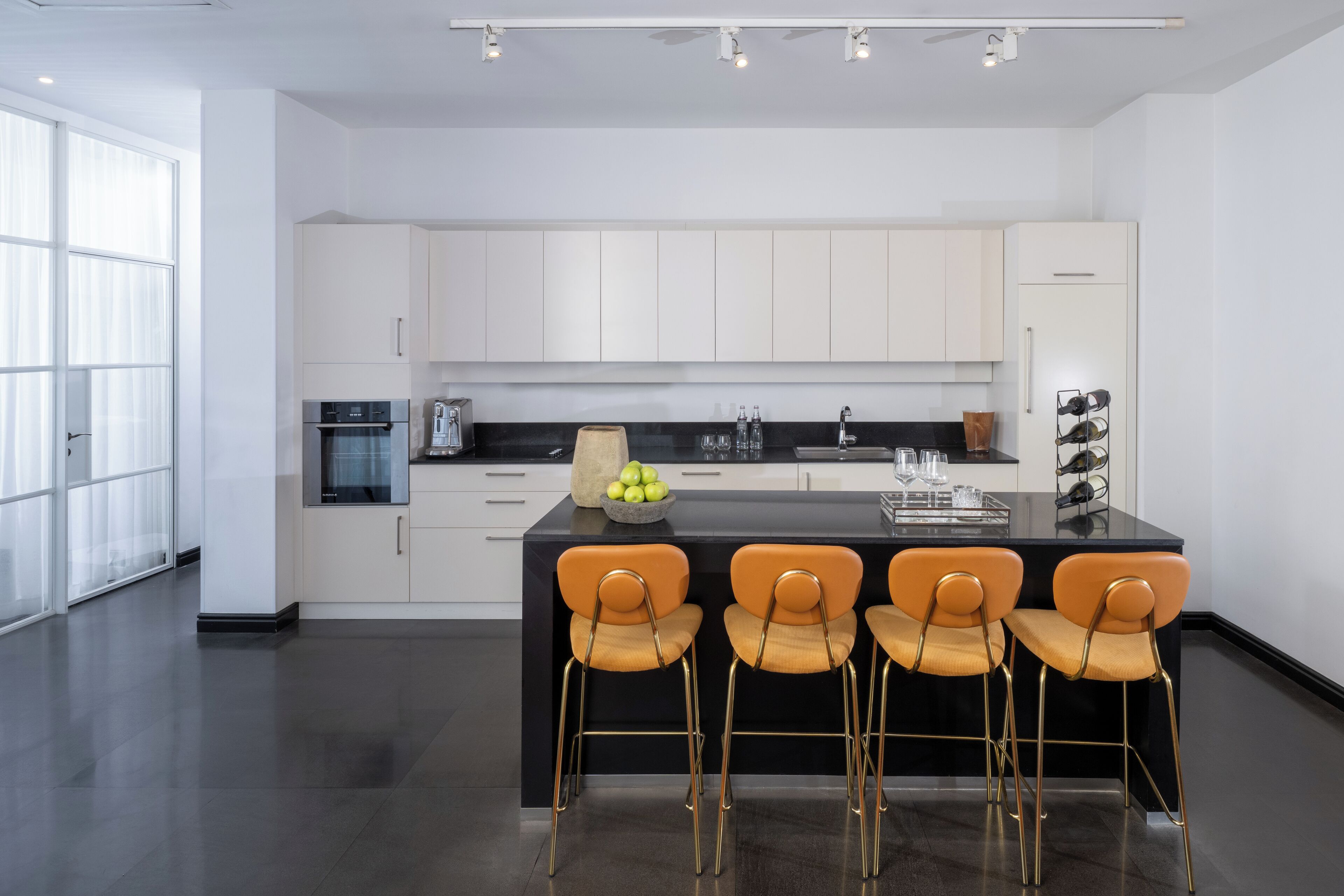 A modern kitchen with white cabinets, a black countertop, and four orange chairs.