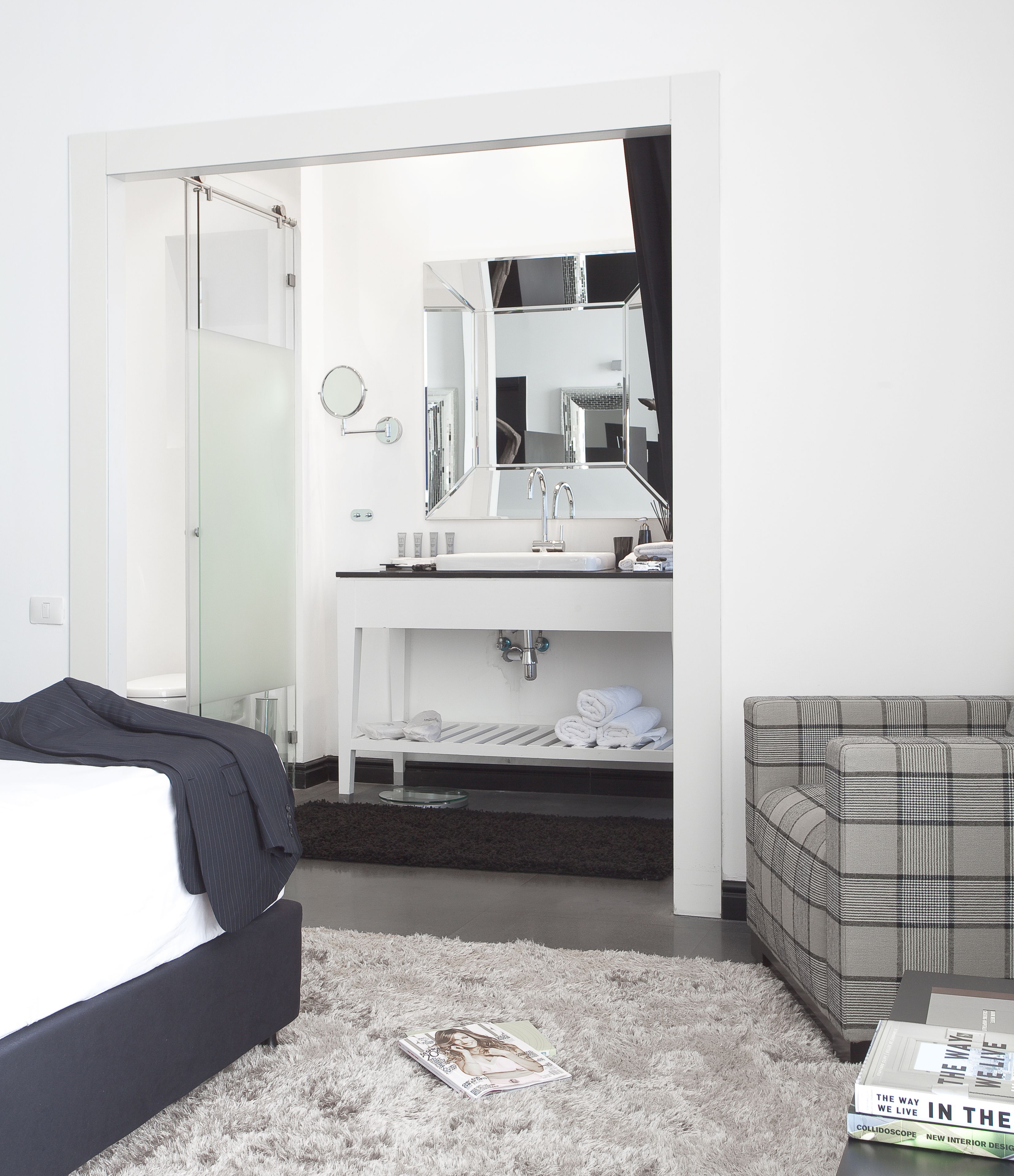 A modern bedroom with a bed, a bedside table, a mirror, a bathroom with a sink and mirror, a grey sofa, and a magazine on the floor.